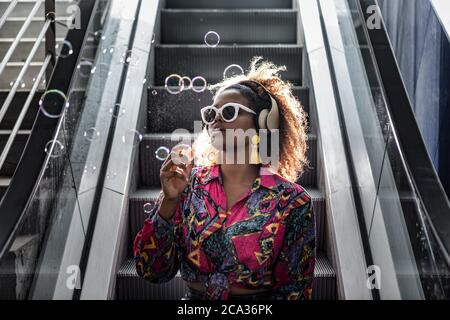 Dama afroamericana en gafas de sol y blusa de colores sentada en escalera en movimiento con boca abierta mientras soplaba burbujas en la parte de atrás iluminada.