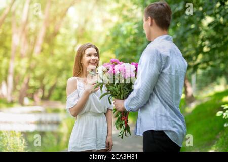 Joven cariñoso sorprendiendo a su novia con ramo de flores en el parque Foto de stock