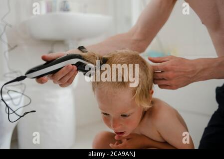 Padre haciendo corte de pelo para su hijo en casa con máquina de corte de pelo en el baño Foto de stock