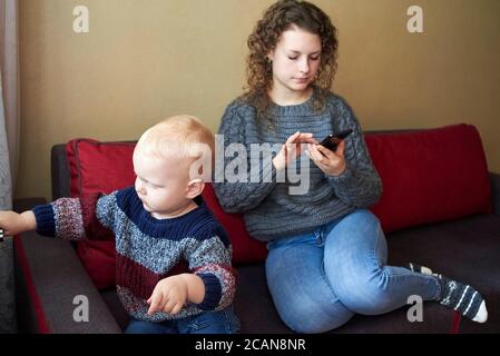 La madre es indiferente a su hijo pequeño, mamá mira el smartphone, el niño necesita atención. El tema de las relaciones padre-hijo. Foto de stock