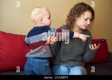 La madre es indiferente a su hijo pequeño, mamá mira el smartphone, el niño necesita atención. El tema de las relaciones padre-hijo. Foto de stock