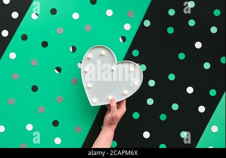 Banner abstracto o plantilla de tarjeta con confeti, lunares. La mano sostiene la caja de luz en forma de corazón. pap en diagonal verde, blanco y negro superpuestos Foto de stock