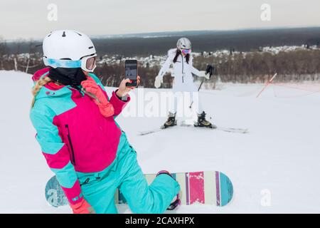 Mujer joven sosteniendo el teléfono inteligente y tomando la foto de su mejor amiga con equipo de esquí, tiempo libre y libre, espacio de copia Foto de stock