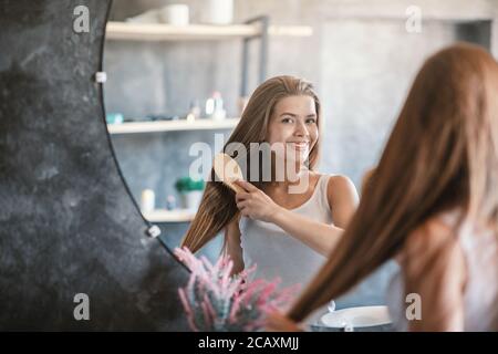 Mujer joven cepillando su hermoso cabello largo con cepillo de madera cerca del espejo interior, copiar espacio Foto de stock