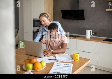 Los jóvenes mestizos pareja casada tener el desayuno a la mañana en la cocina y el uso de ordenador portátil. Las compras en línea.