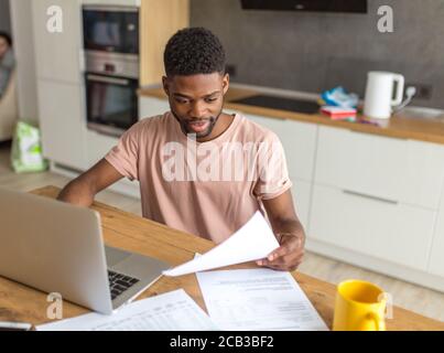 Apuesto hombre de piel oscura habiendo meditado y grave expresión facial mientras revisa sus finanzas y pagar facturas en línea, sentado en una mesa con docu