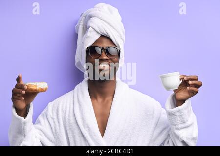 Hombre atractivo y guapo en toalla y albornoz sosteniendo una taza de té y donut, emocional. Niño fresco en anteojos posando con donuts aislados sobre púrpura Foto de stock