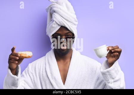 Hombre atractivo y guapo en toalla y albornoz sosteniendo una taza de té y donut, emocional. Niño fresco en anteojos posando con donuts aislados sobre púrpura Foto de stock