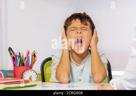 Niño autista grita y cierra los oídos con dolor durante ABA terapia de desarrollo sentado junto a la mesa en clase Foto de stock