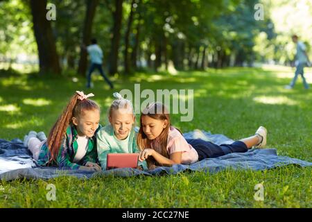 Retrato completo de tres chicas adolescentes usando una tableta digital mientras se tumban sobre hierba verde en el parque al aire libre iluminado por la luz del sol, copiar espacio