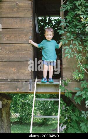 Un niño pequeño está parado en las escaleras de la casa del árbol en el jardín verde. Horario de verano