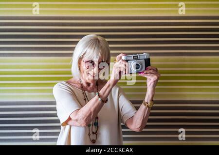 Elegante mujer mayor con gafas de sol fotografiando con cámara contra pared en casa