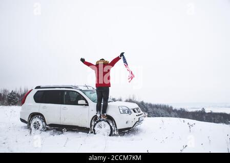 Hombre de pie en la cima de la colina con la bandera de EE.UU. en sus manos cerca de suv coche buscando hermosa vista de invierno