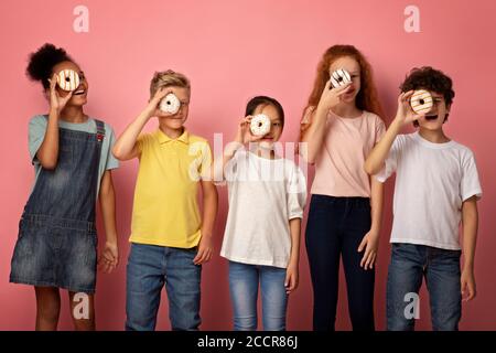 Escolares diversos sosteniendo buñuelos deliciosos delante de sus caras sobre fondo rosa Foto de stock