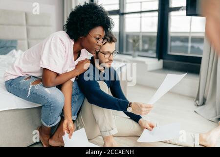 Interracial pareja de jóvenes recién casados sentarse juntos en el piso de sus apartamentos alquilados, ocupado por componer informe financiero para servicio tributario, utilizando p