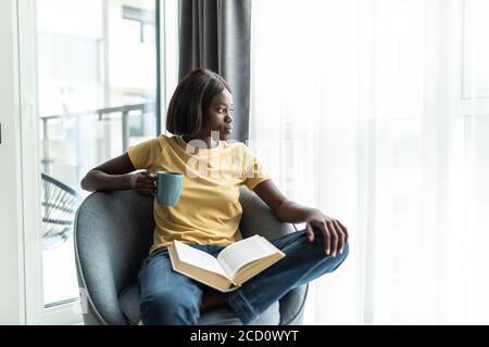 Ocio en casa. Feliz niña negra leyendo libro y bebiendo café, sentado en una silla de mimbre contra la ventana en la sala de estar, espacio de copia