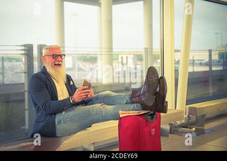 Hombre de moda mayor usando un teléfono inteligente dentro del aeropuerto internacional. Retrato de un hombre de negocios mayor caucásico. Adulto sonriente barbudo elegante éxito bu