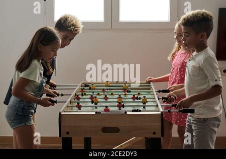 Un grupo de niños multiculturales y lindos están en el interior pasar tiempo de juego con amigos jugando al futbolín juntos. Versión de mesa de los amantes del fútbol.