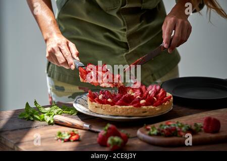 Vista de la cosecha de la cocinera anónima corte de fresa casera fresca pastel mientras está de pie en la cocina en la mesa de madera