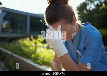 pandemia de coronavirus. mujer médica moderna estresada en uniforme con estetoscopio y máscara médica sentada al aire libre cerca del hospital. Foto de stock