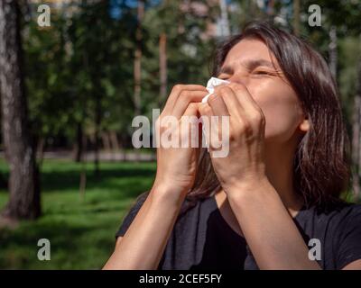 Mujer con ojos cerrados y gry que sufre de polen de pasto estornuda en papel en un día soleado en el parque. Concepto de funcionamiento de la nariz alérgica. Foto de stock