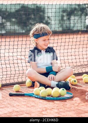 Niño pequeño con raqueta de tenis y sentado cerca de la red de tenis entre las bolas esparciendo en la cancha en el día soleado Foto de stock
