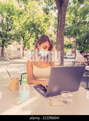 Mujer joven usando máscara protectora y desinfectante de manos mientras trabaja remotamente en un ordenador portátil en una cafetería al aire libre. La Nueva normal an Foto de stock