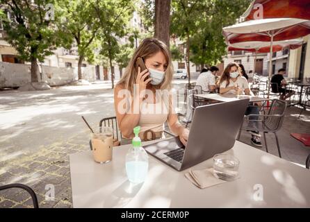 Mujer joven usando máscara protectora y desinfectante de manos mientras trabaja remotamente en un ordenador portátil en una cafetería al aire libre. La Nueva normal an Foto de stock