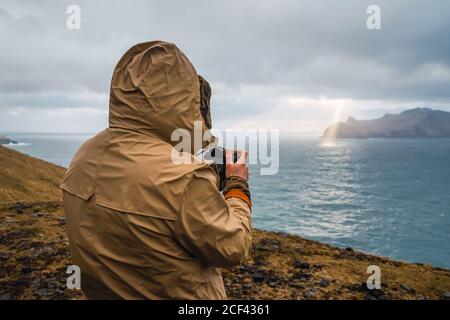 Vista posterior de hombre anónimo en ropa de invierno utilizando un Cámara fotográfica al aire libre en el paisaje de las Islas Feroe