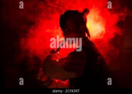 Rebelde mujer en chaqueta informal con piercing y moderno peinado  sosteniendo murciélago entre la luz roja de colores y el vapor Fotografía  de stock - Alamy