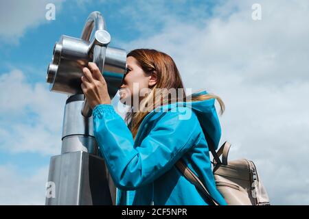 mujer rubia anónima en abrigo azul mirando a través de la moneda de la calle operado binocular mientras está de pie sobre pavimento de madera cerca de la valla de concreto en el día soleado Foto de stock