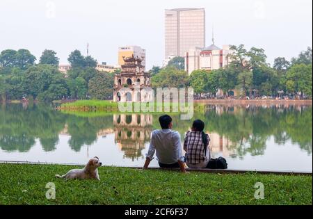 Amigos hablando a lo largo del lago Hoan Kiem con la pagoda tortuga y modernos edificios en el fondo en Hanoi, Vietnam. Foto de stock