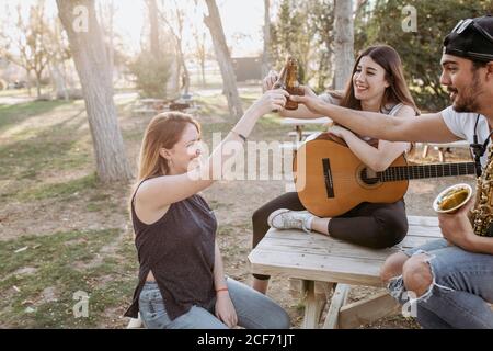 Grupo de jóvenes amigos con instrumentos musicales sonriendo y sonriendo botellas de cerveza mientras pasan tiempo juntos en el parque