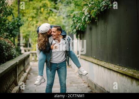 Mujer contenta con ropa informal sentada en la espalda y besándose feliz hombre caminando en un pequeño callejón con plantas verdes fondo borroso