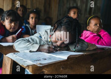 Madagascar - 6 DE JULIO de 2019: Niños africanos atentos escuchando y escribiendo en cuadernos mientras se sentaban en los escritorios de la escuela rural
