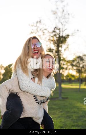 Rubia chica piggyback montar en su amigo. Bromeando y sonriendo los felicita. Luz de fondo en un día soleado en el parque. Foto de stock