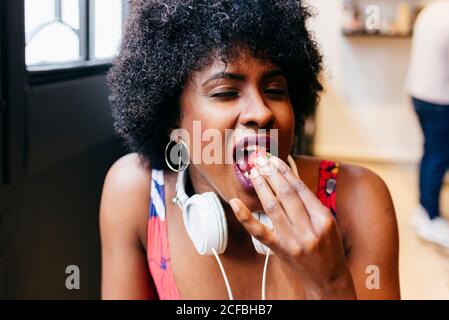Mujer negra bastante joven con auriculares en el cuello tomando el desayuno en un pequeño café y disfrutando de una deliciosa fresa fresca
