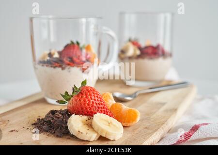 Tazas de cristal de delicioso yogur fresco decorado con fresa chocolate y trozos de plátano y mandarina colocados en madera tabla de cortar sobre fondo gris