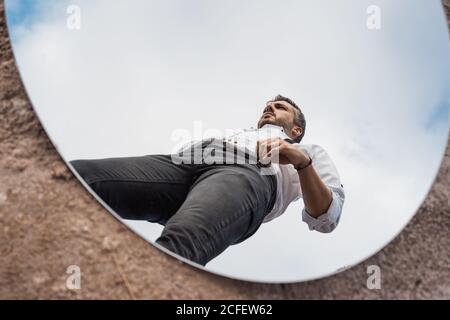 Reflejo de bajo ángulo del hombre soñador en camisa y tirantes de pie sobre el cielo azul en el espejo ovalado en tierra polvorienta