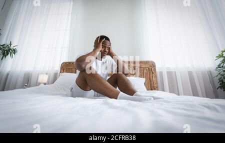 Estresado Hombre del Milenio Negro sentado en la cama tocando la cabeza Interior Foto de stock