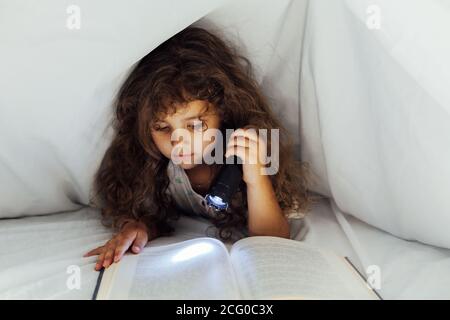 Hermosa niña lee un libro con una linterna debajo una manta