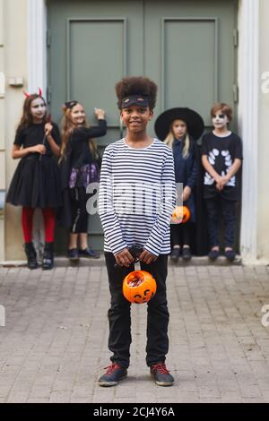 Retrato vertical de toda la longitud de un grupo multiétnico de niños que visten trajes de Halloween mirando la cámara mientras se trampean o tratan juntos, se centran en el niño afroamericano en primer plano