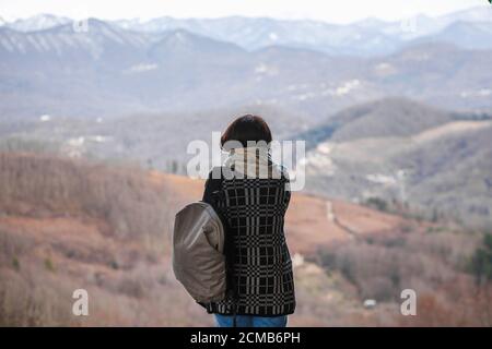 Una mujer con una mochila mira hacia las montañas en una tarde fría a finales de otoño. Foto de stock