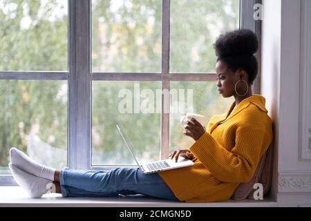 Mujer afroamericana enfocada estudiante con afro peinado llevar cárdigan amarillo, sentado en el alféizar de la ventana, trabajando haciendo trabajo remoto en el portátil, preparando
