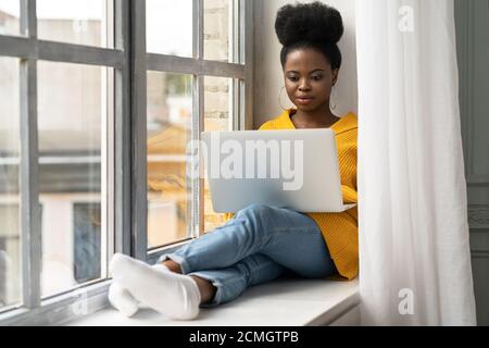 Mujer afroamericana estudiante con afro peinado llevar cárdigan amarillo, sentado en el alféizar de la ventana, trabajando haciendo trabajo remoto en el portátil, aprendiendo usando onl