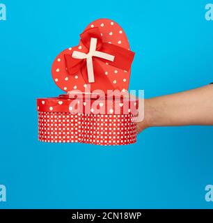 la mano sostiene una caja de cartón roja en forma de corazón con cinta de seda sobre un fondo azul Foto de stock
