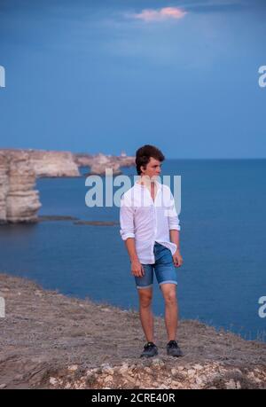 Un joven está de pie en una playa rocosa al atardecer.