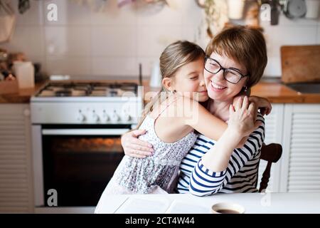 Feliz madre está abrazando a su hija en la acogedora cocina de casa. La mujer y la niña linda están sonriendo.