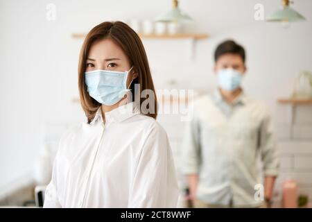 retrato de una joven mujer asiática con máscara mirando cámara con esposa en el fondo