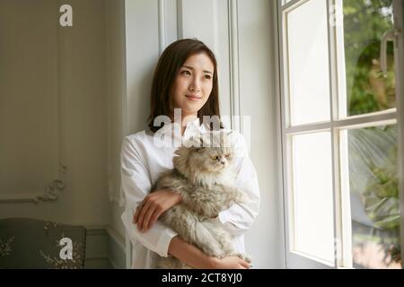 hermosa joven asiática de pie junto a la ventana en la casa de celebración un gato en los brazos mirando serena y contento